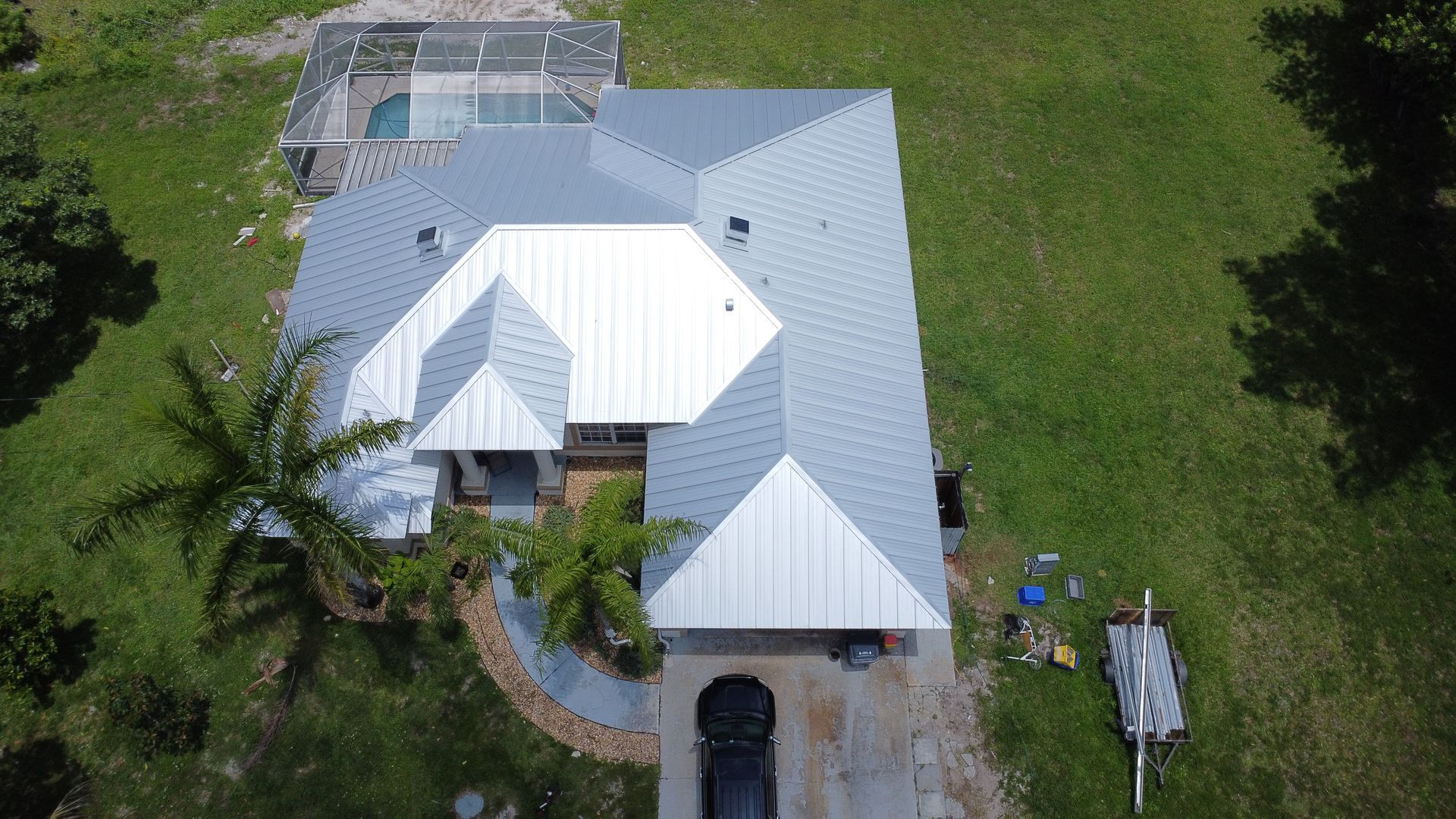 Aerial view of a white-roofed house surrounded by green lawn and trees, with a driveway at the front.