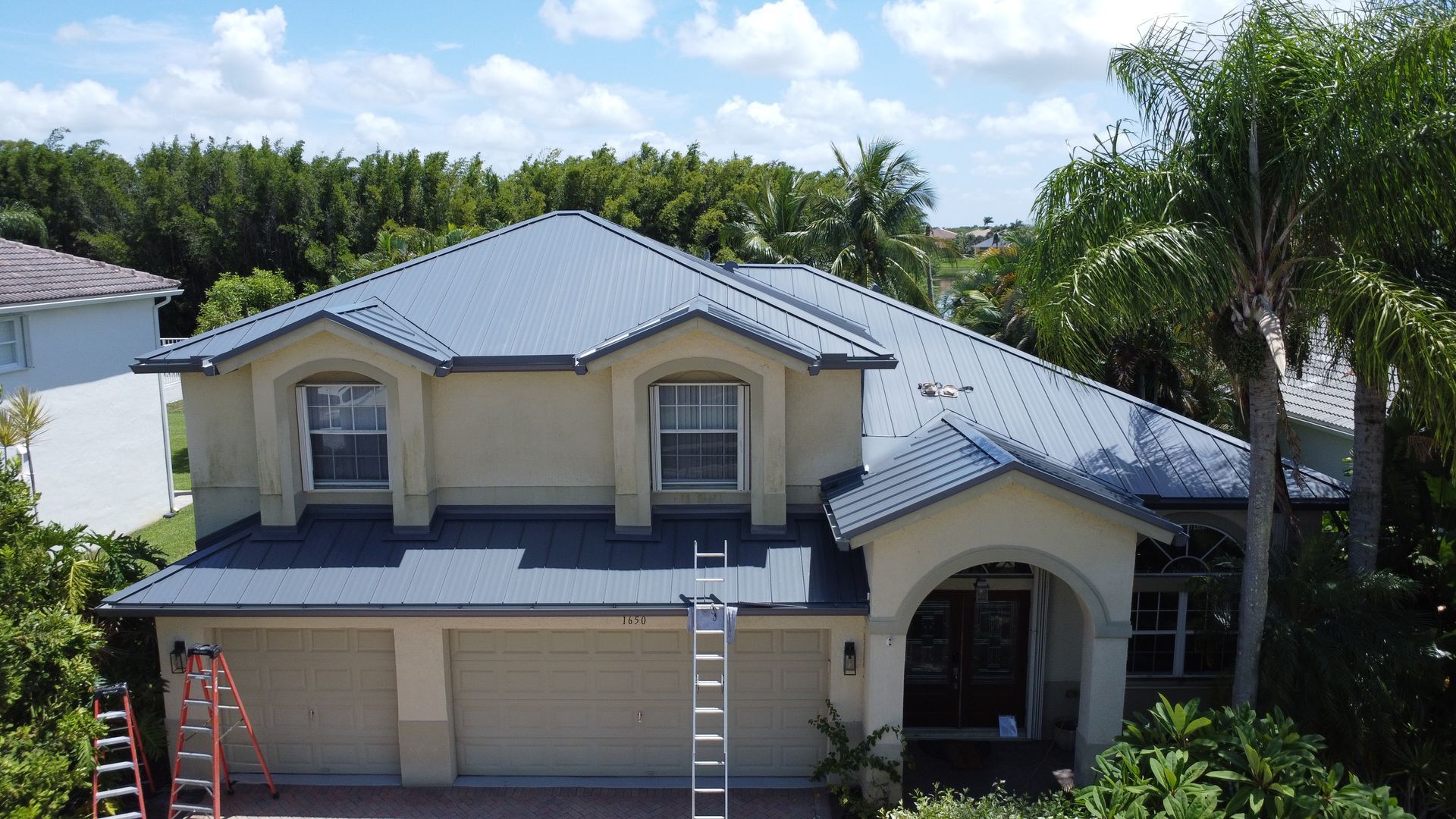 Two-story suburban house with blue roof, ladders in the driveway, and palm trees around it