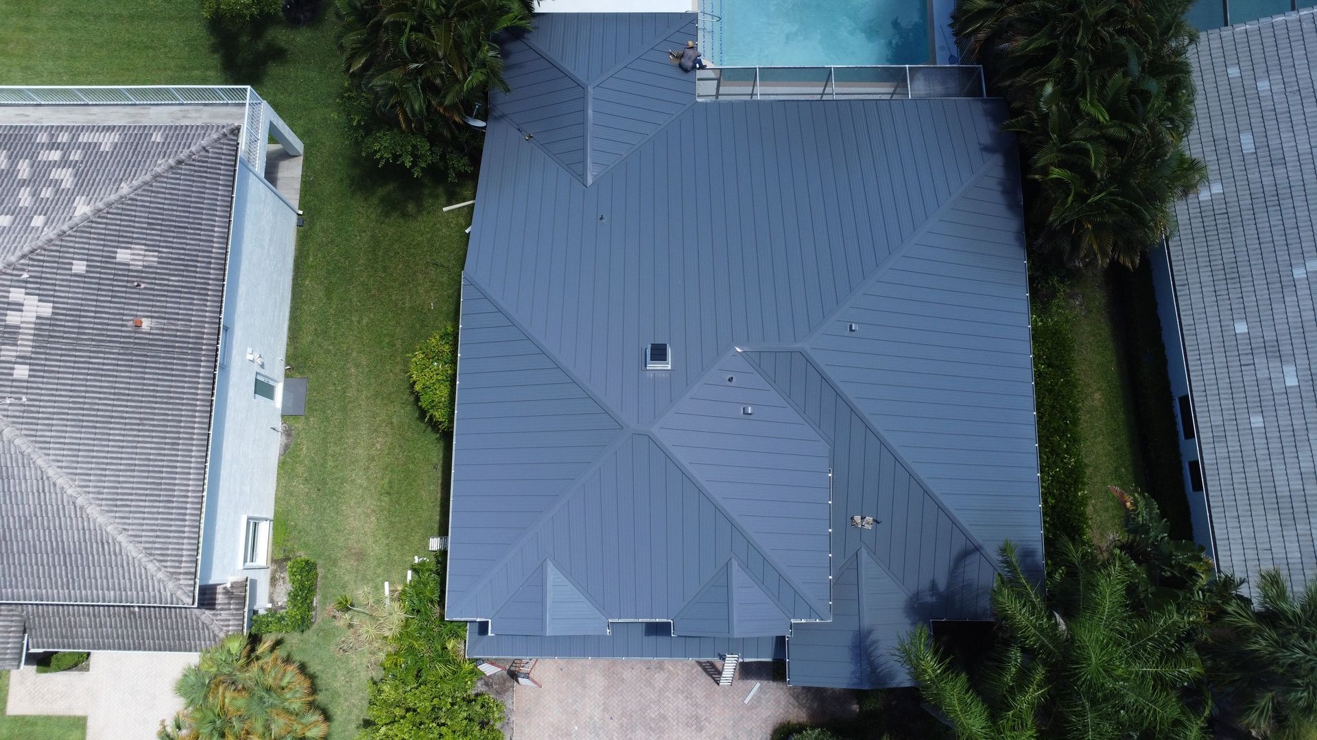 Aerial view of a gray-roofed house with a pool, surrounded by trees and neighboring homes.