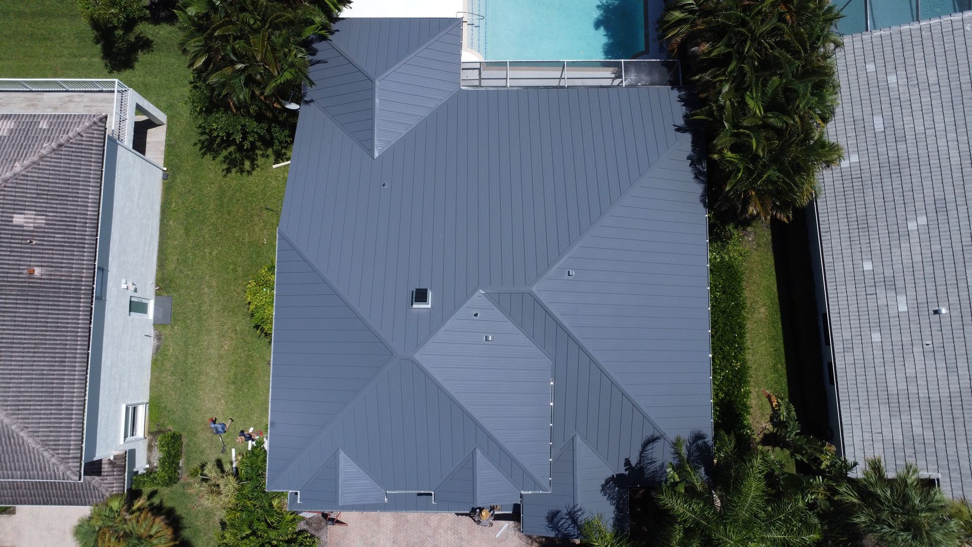 Aerial view of a gray-roofed house with a backyard pool, bordered by trees and neighboring homes.