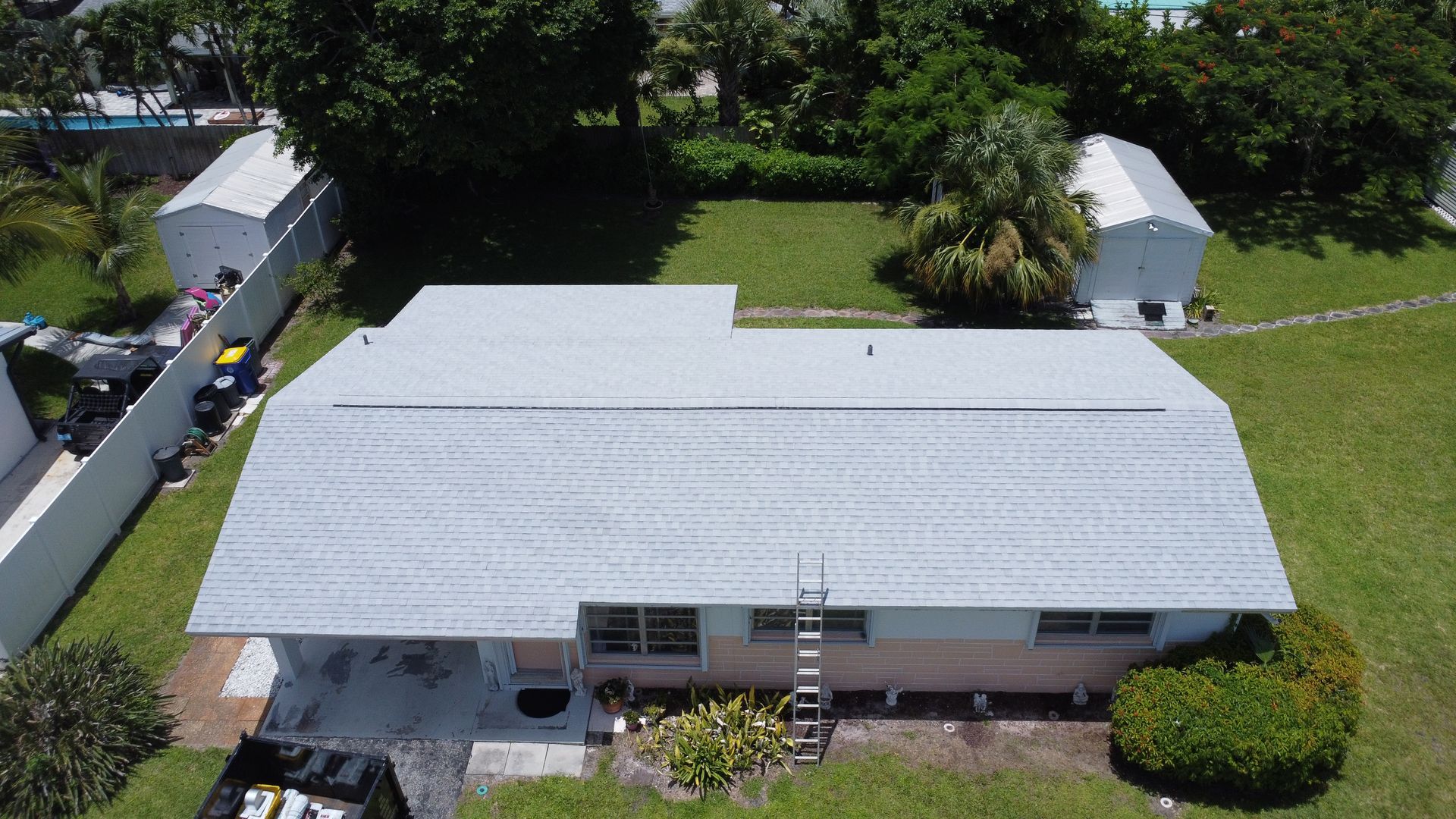 Aerial view of a white-roofed house with a ladder, surrounded by grass and trees.