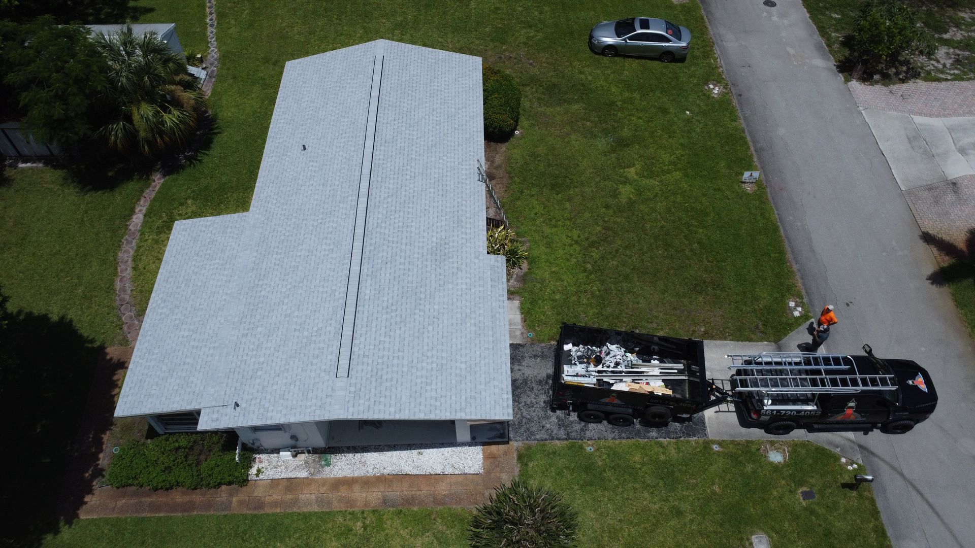 Aerial view of a roof being repaired, with a worker and truck beside a house on a grassy lot.