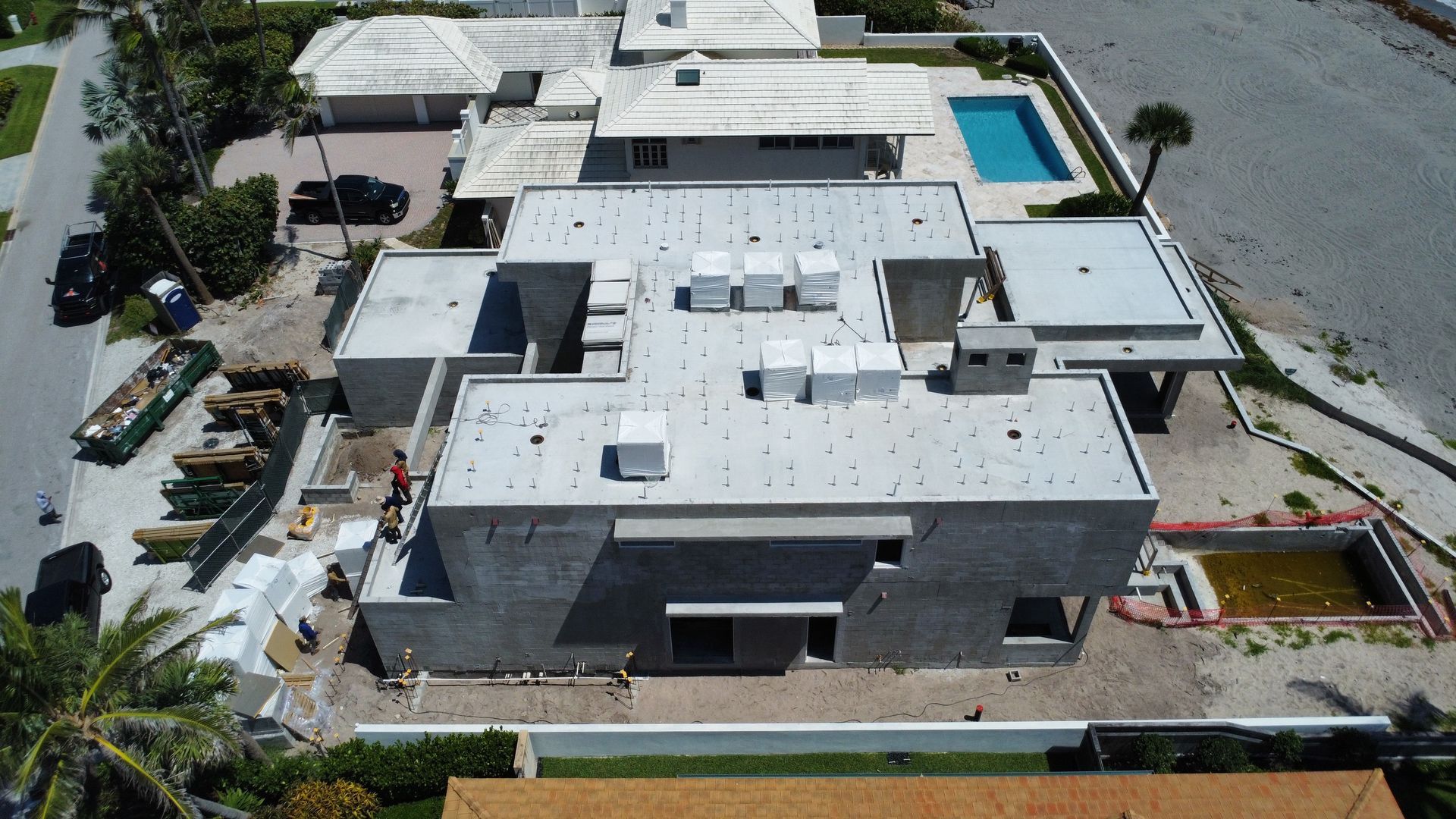 Aerial view of a large flat-roofed white building with parking, palm trees, and a swimming pool nearby.