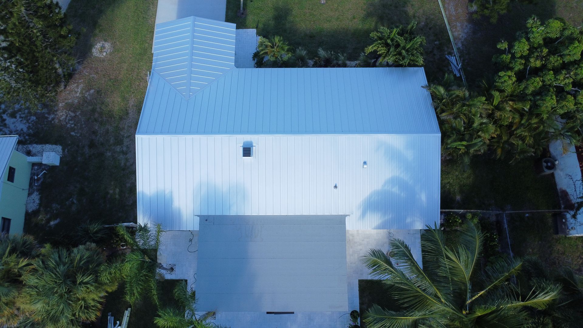 Aerial view of a white house with a light blue roof surrounded by dense green trees.