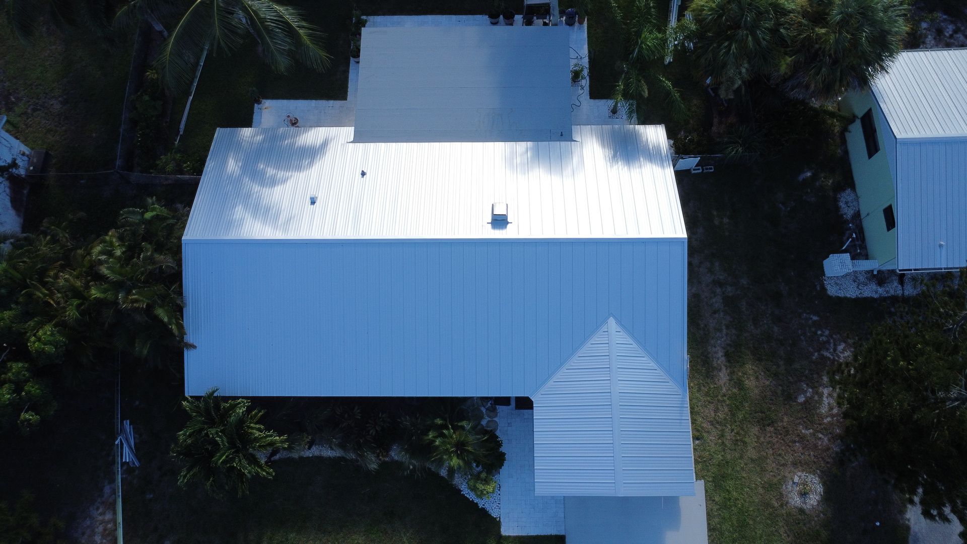 Aerial view of a white-roofed house surrounded by trees, with a small side structure and driveway.