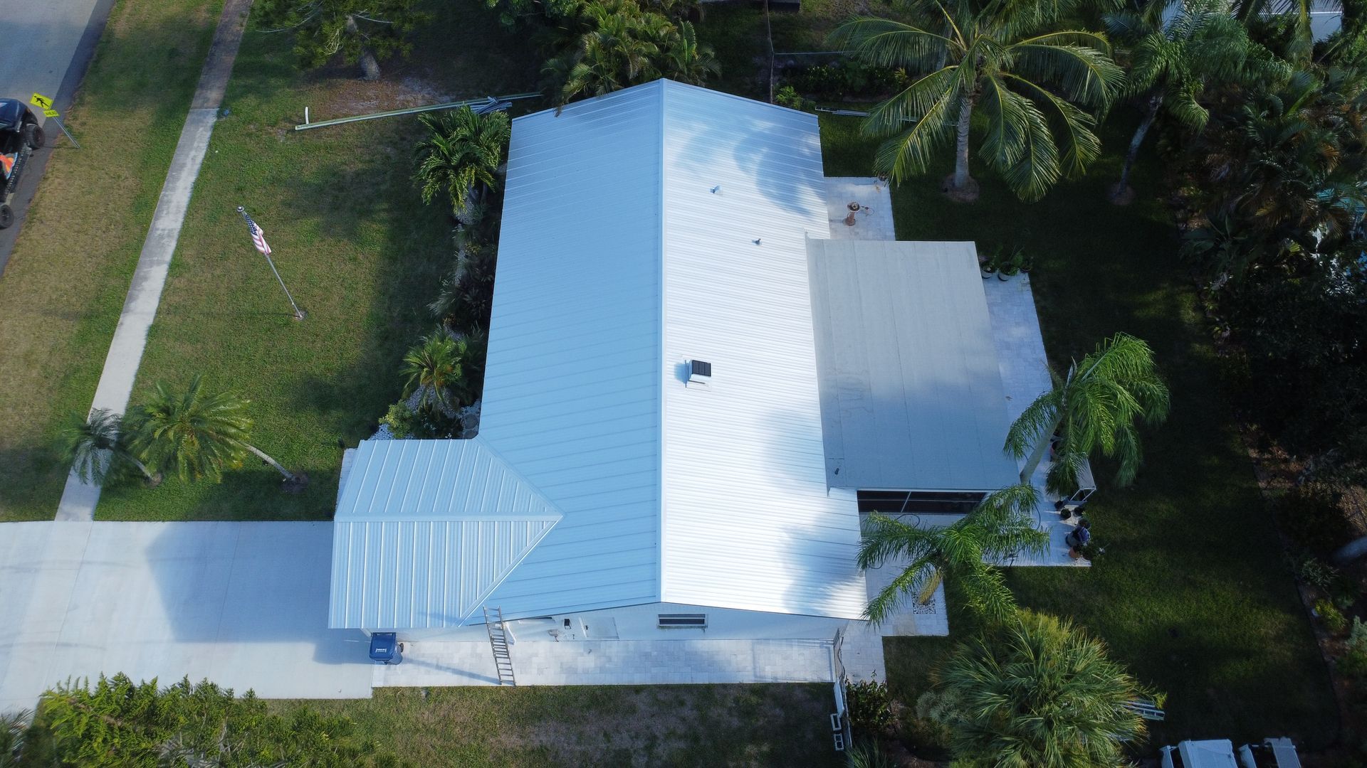 Aerial view of a white-roofed house with a driveway, surrounded by green trees and lawn.