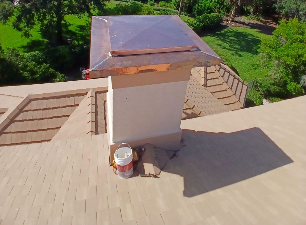 Rooftop with beige shingles and a central vent stack under sunlight, surrounded by green trees.