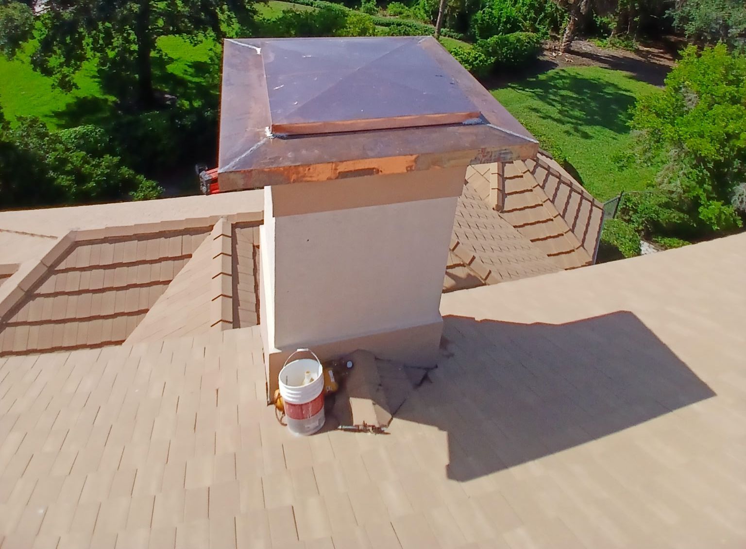 Rooftop with beige shingles and a central vent stack under sunlight, surrounded by green trees.