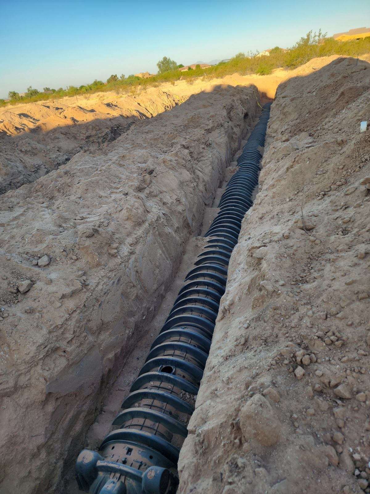 Black corrugated pipe laid in a deep dirt trench under a bright sky
