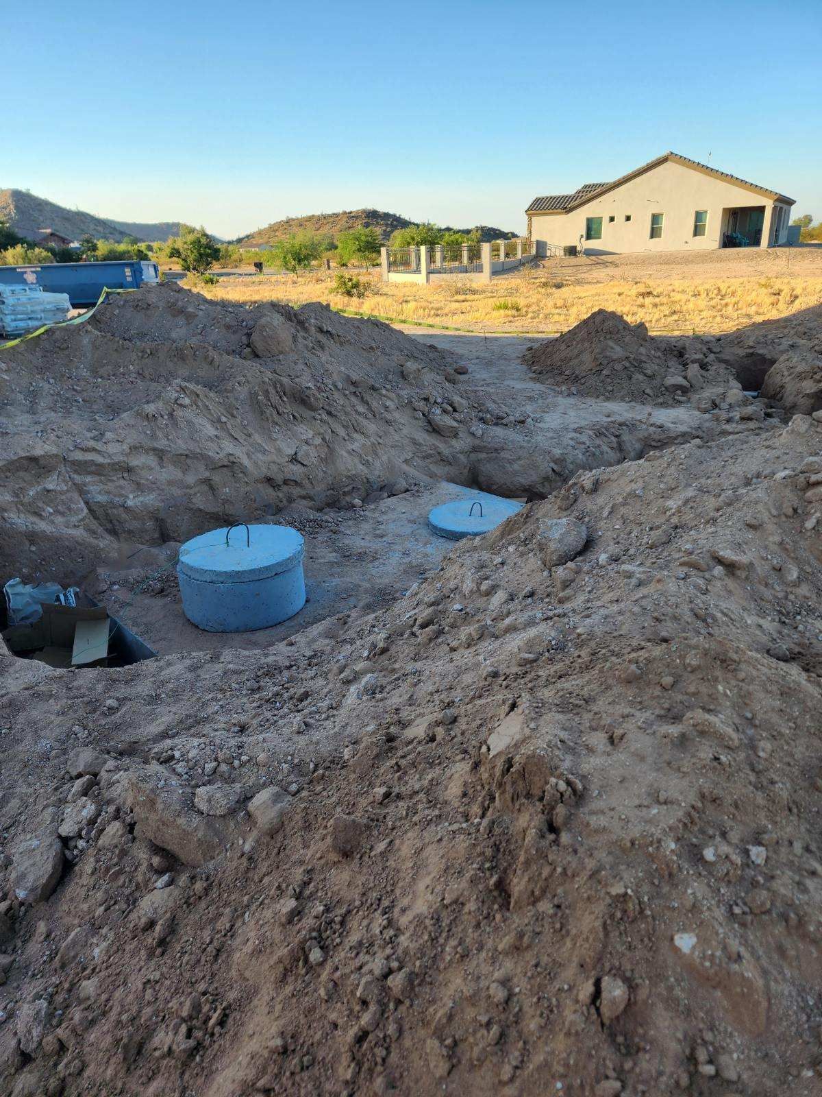 Construction site with dirt trenches, blue utility covers, and a house in the background