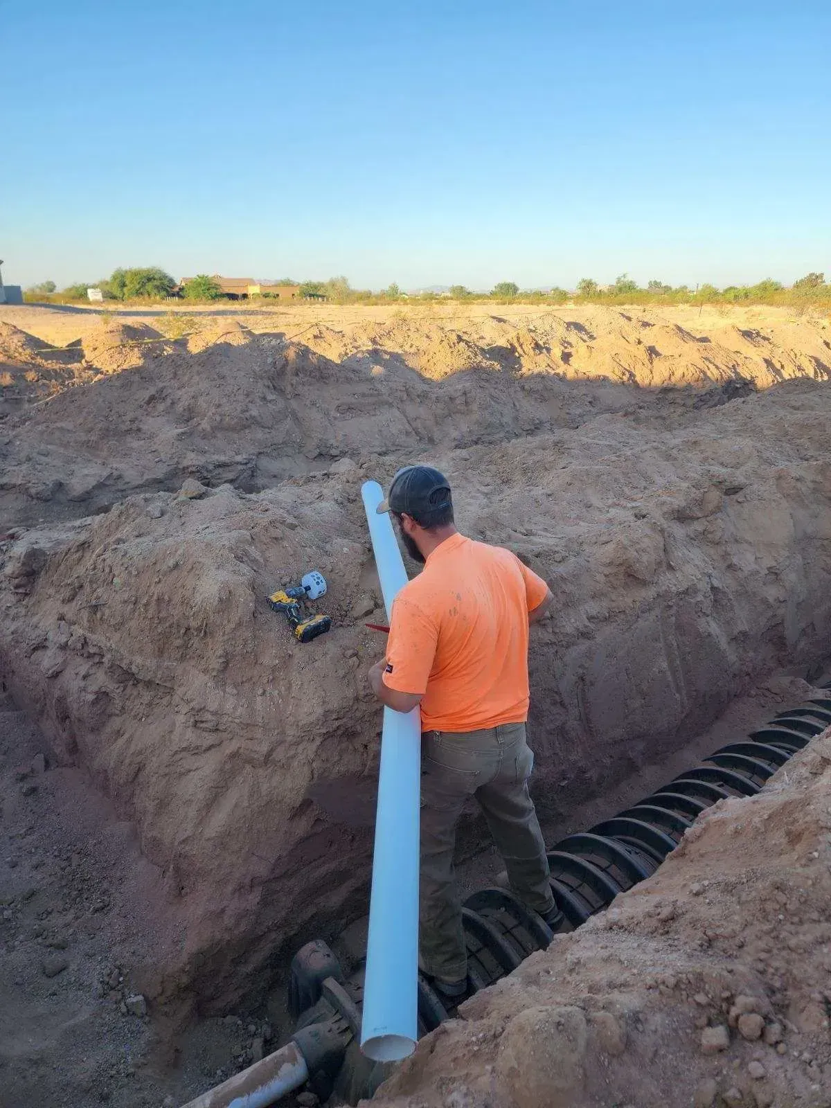 Worker in orange shirt installing a large pipe in a trench at a construction site
