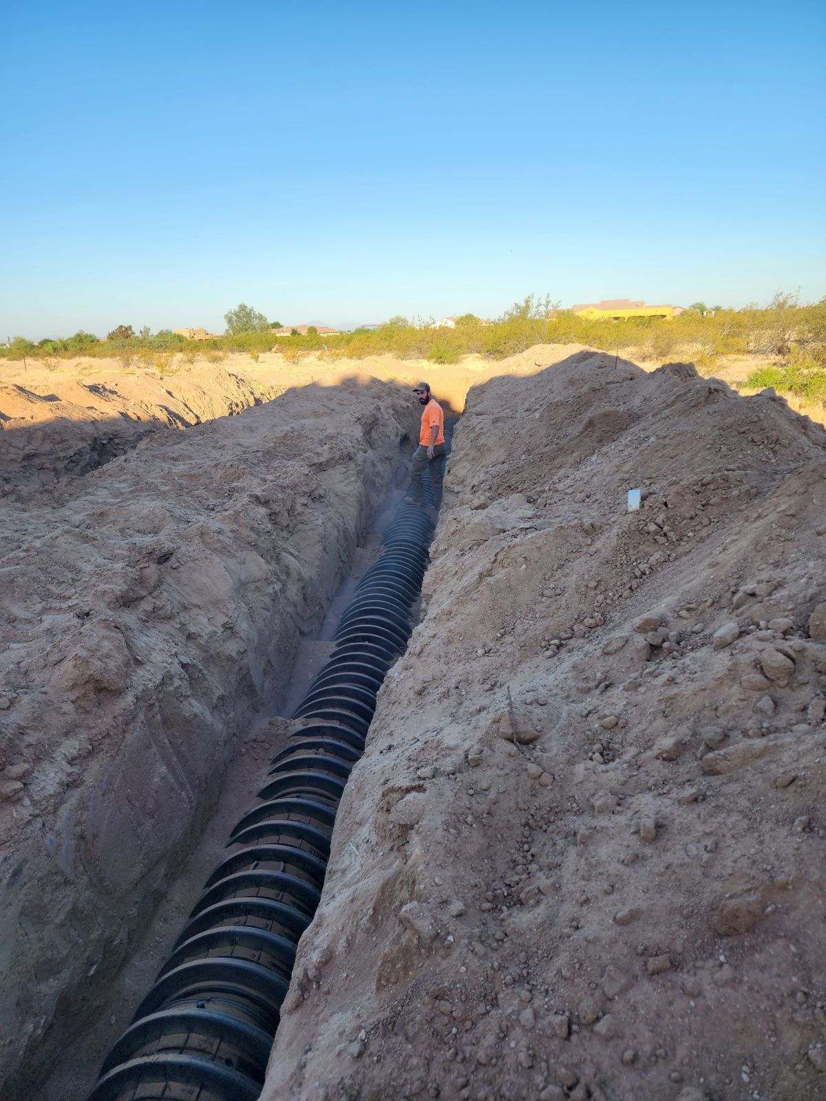 Trench with black corrugated pipe laid in the dirt beneath a blue sky.
