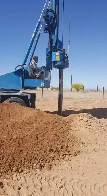Blue drilling rig boring into dirt in a dry field, with a large soil mound in the foreground