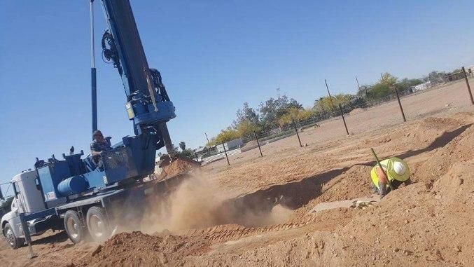 Drilling rig at a dusty construction site, with a worker in a hard hat crouched in a trench.