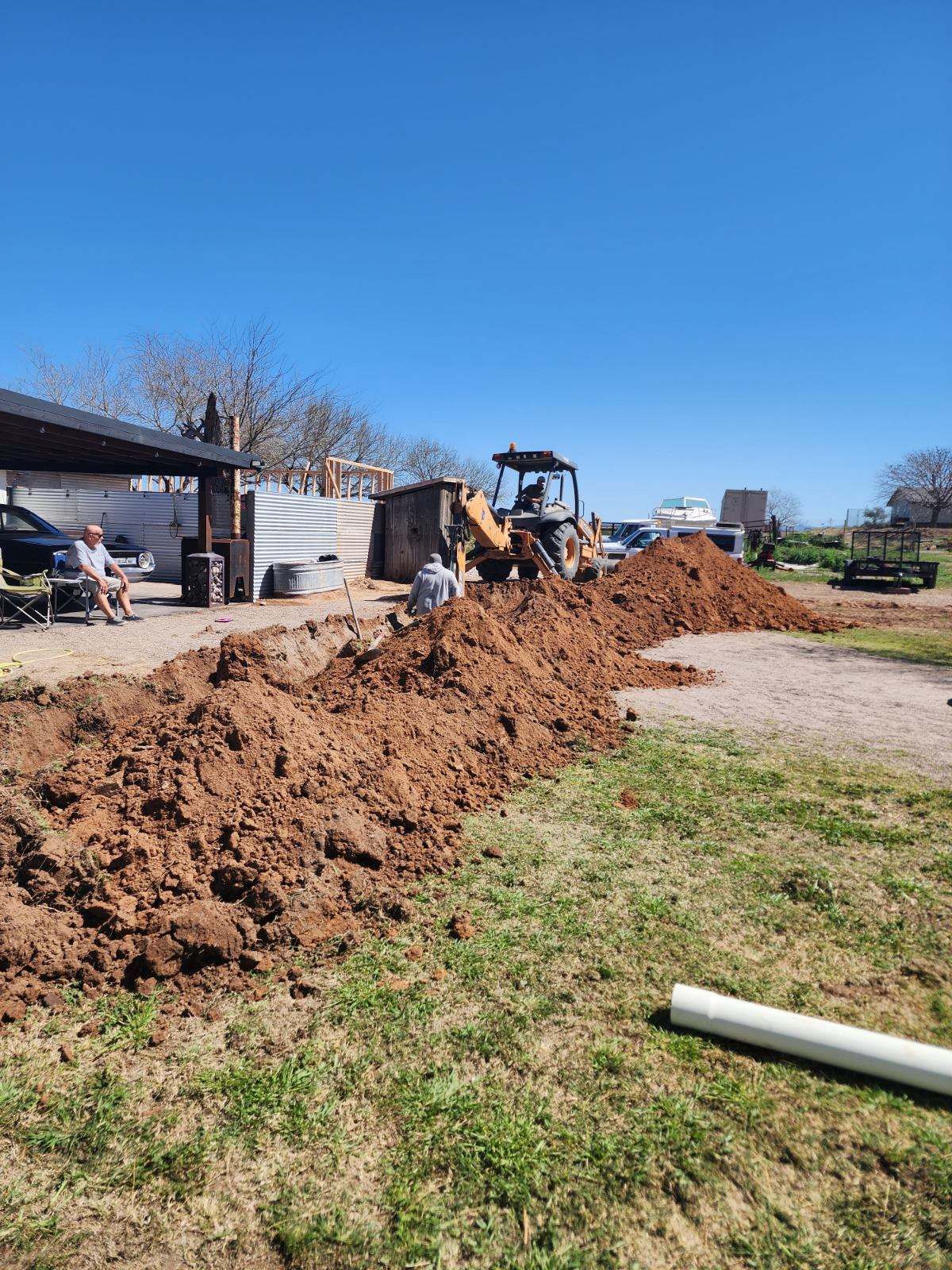 Excavator beside a long dirt trench in a rural yard near a house on a clear blue day