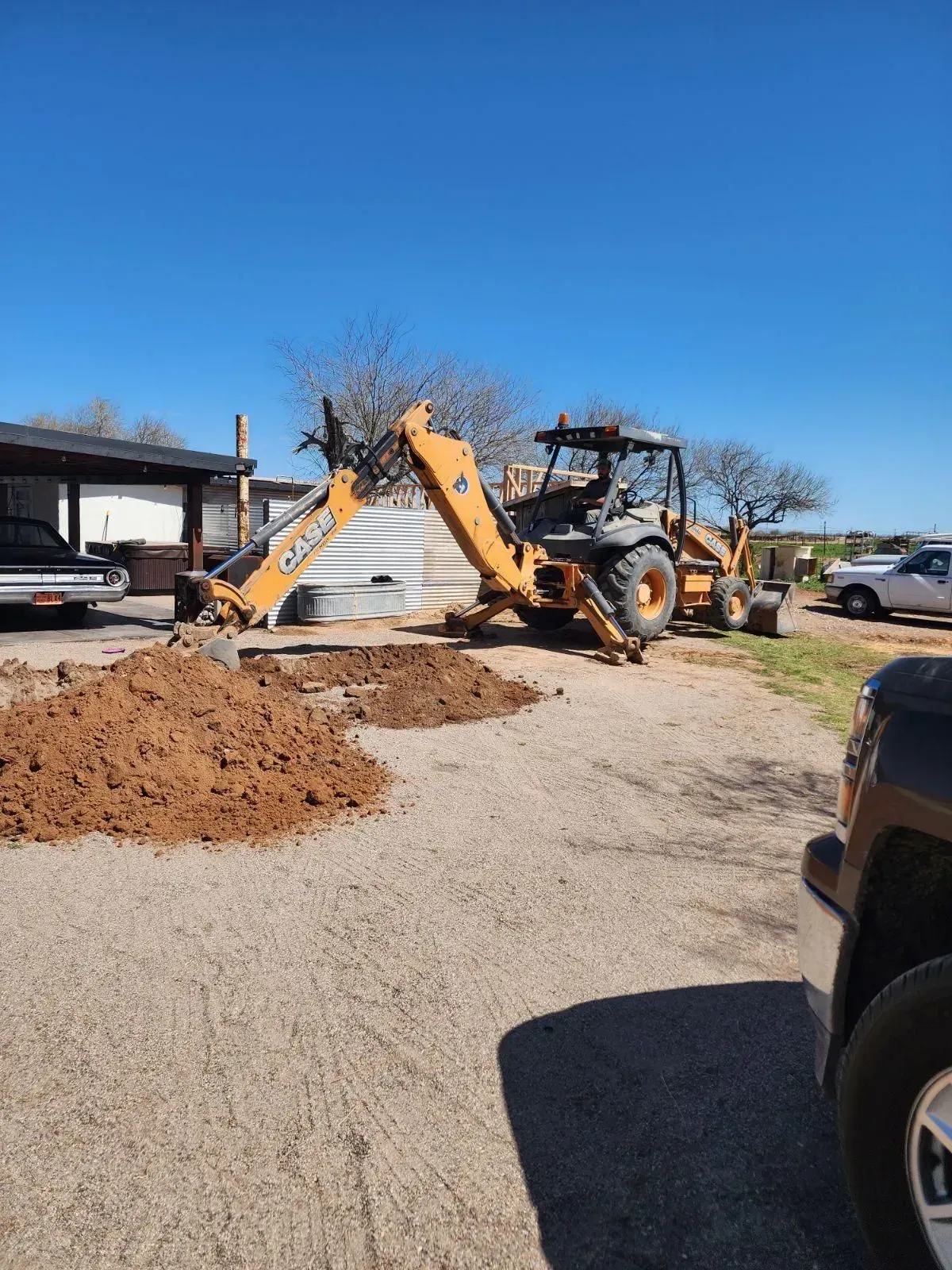 Construction equipment moving dirt in a gravel driveway beside a house under a clear blue sky