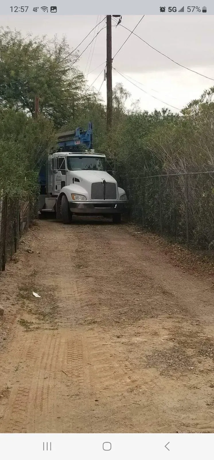 White truck on a narrow dirt road, carrying equipment under trees and power lines.