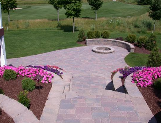 Paved garden patio with curved stone seating, pink flowers, and a circular fire pit overlooking a lawn