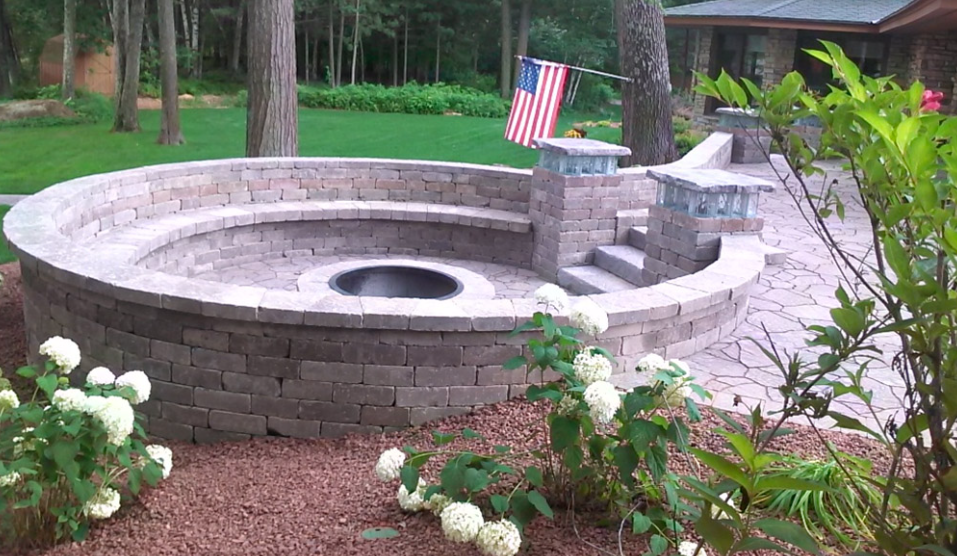 Backyard stone fire pit with curved seating wall, surrounded by flowers and trees, with a small U.S. flag nearby