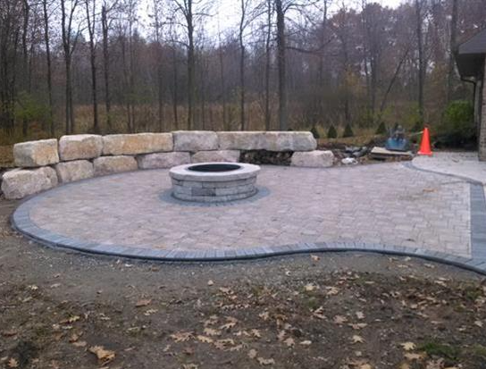 Stone patio with circular fire pit and low wall benches in a wooded yard.