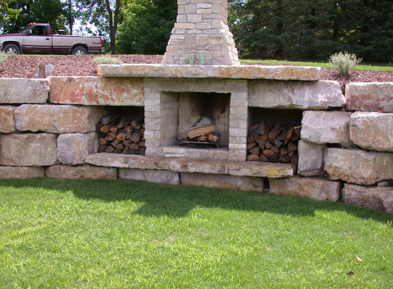 Stone outdoor fireplace built into a low stone wall on a grassy lawn, with a chimney and stacked firewood.