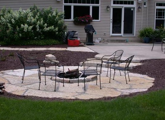 Backyard patio with a stone fire pit surrounded by metal chairs on a lawn