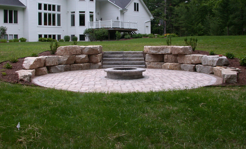Stone patio fire pit with curved rock benches in a grassy yard, in front of a white house