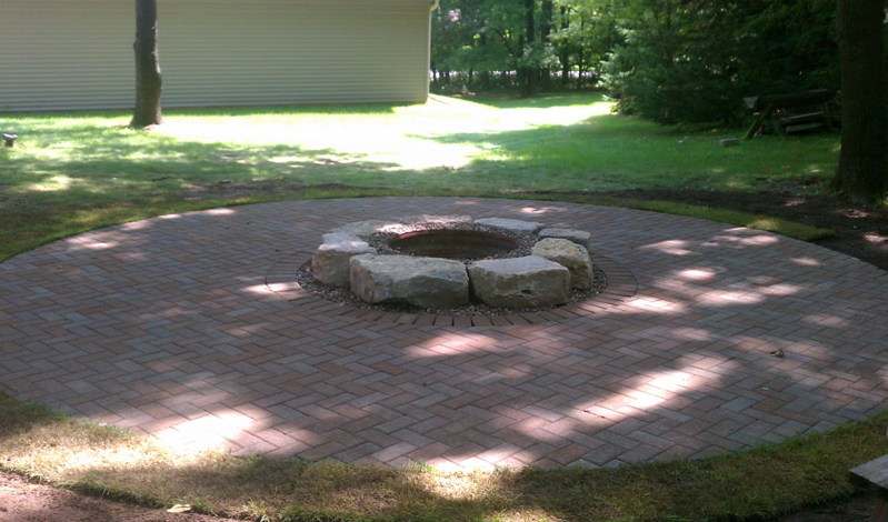 Brick patio with a stone fire pit in a shaded backyard garden area.