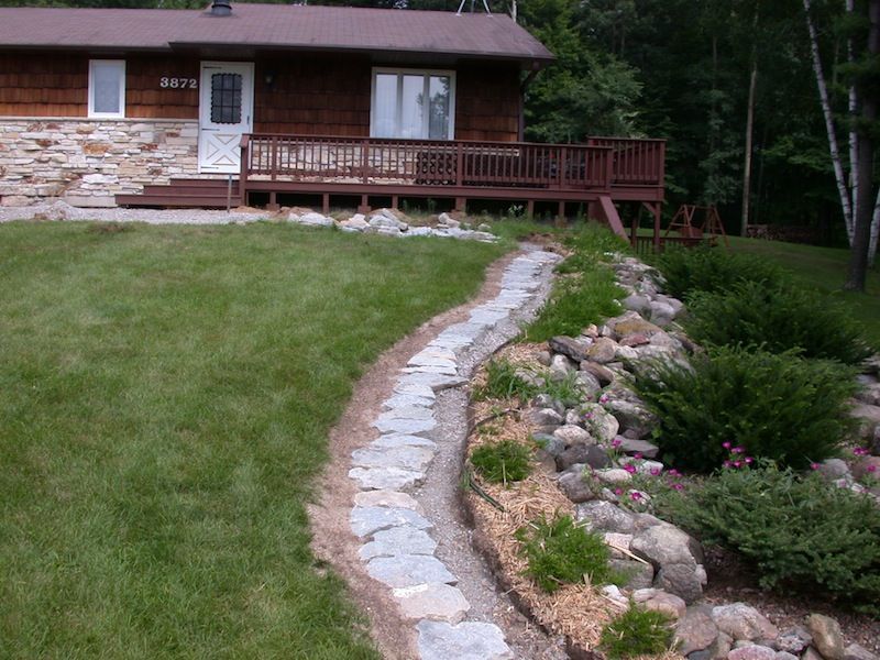 House with wooden deck and stone-edged garden path beside a sloped lawn