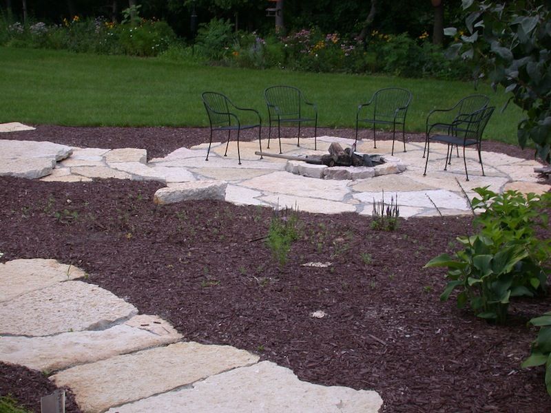 Stone patio with a circular fire pit and metal chairs in a garden, bordered by mulch and greenery