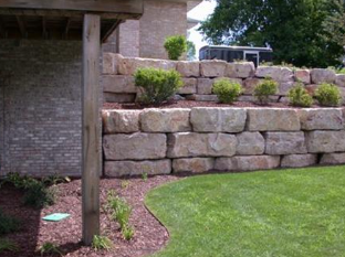 Stone retaining wall with shrubs along a curved green lawn beside a brick house