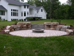 Stone fire pit with curved bench seating in a grassy backyard beside a white house