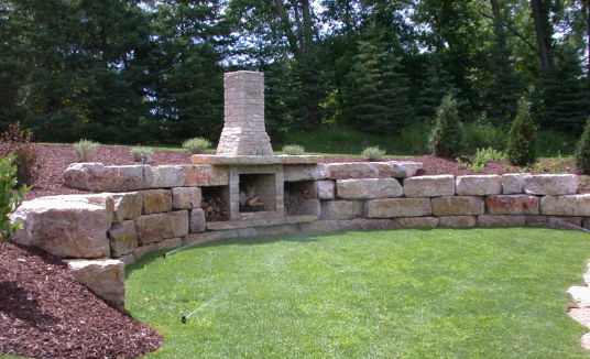 Curved stone retaining wall with built-in brick fireplace beside a green lawn and trees