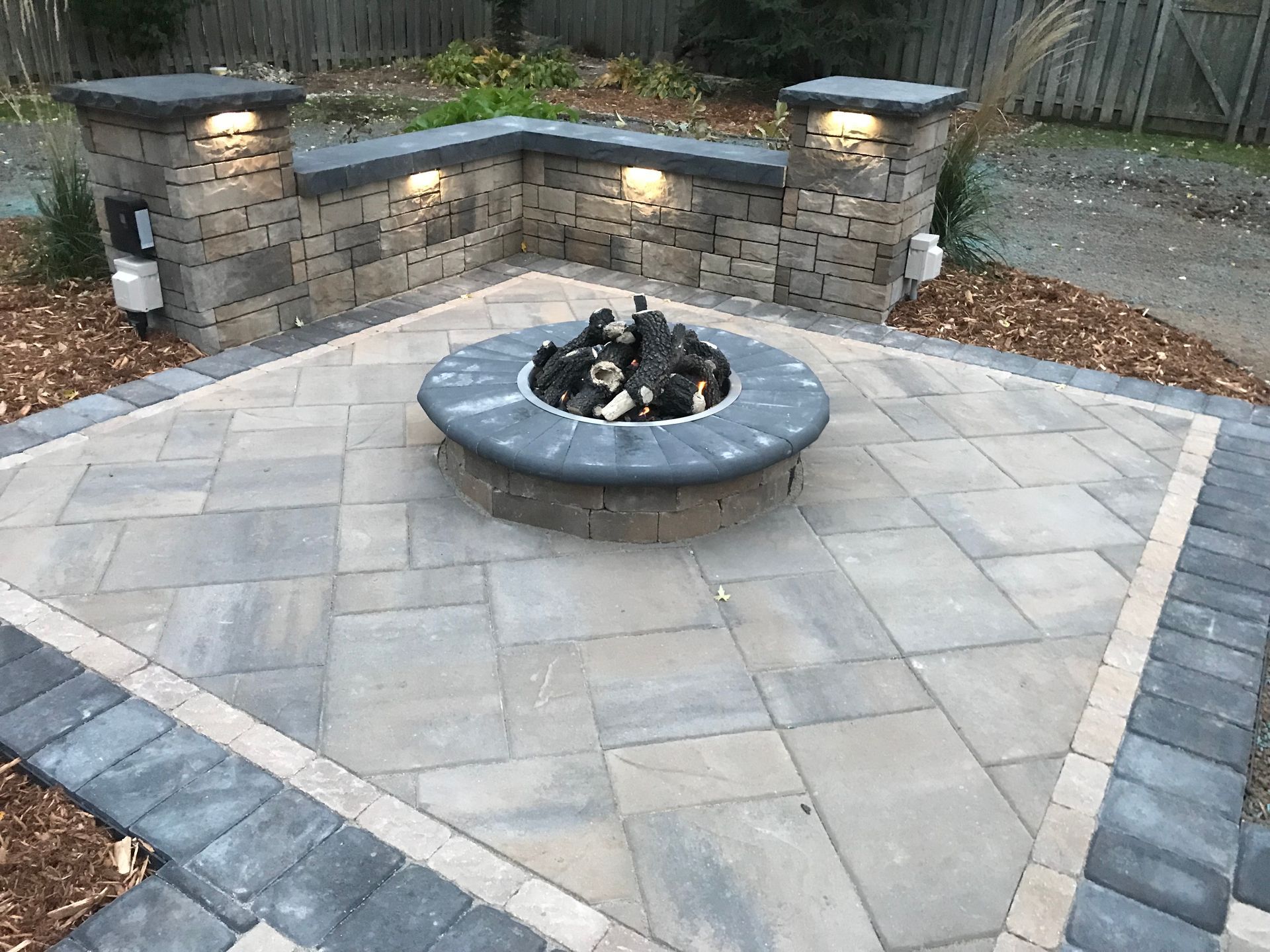 Stone patio with a circular fire pit and built-in lit seating wall at dusk.
