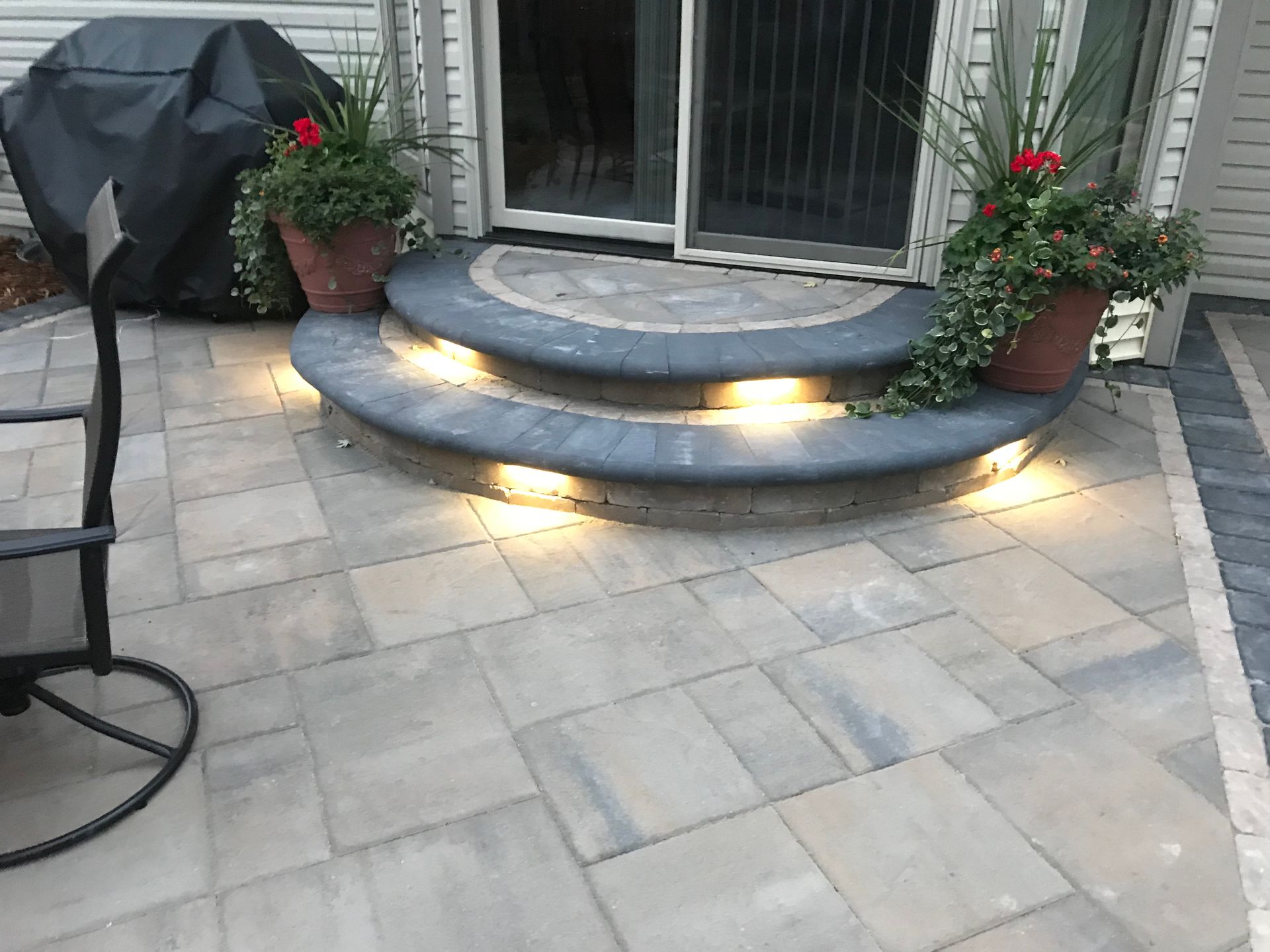 Stone patio steps with warm lights, potted flowers, and a covered grill beside sliding doors