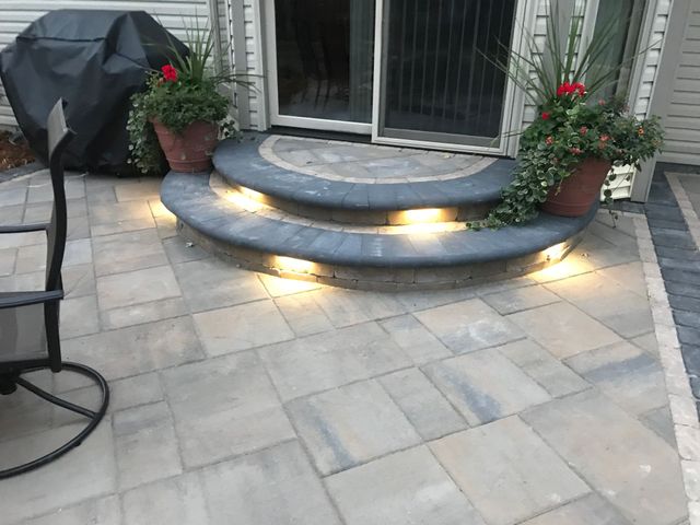 Patio steps with warm lights, potted flowers, stone pavers, and a covered grill beside sliding doors