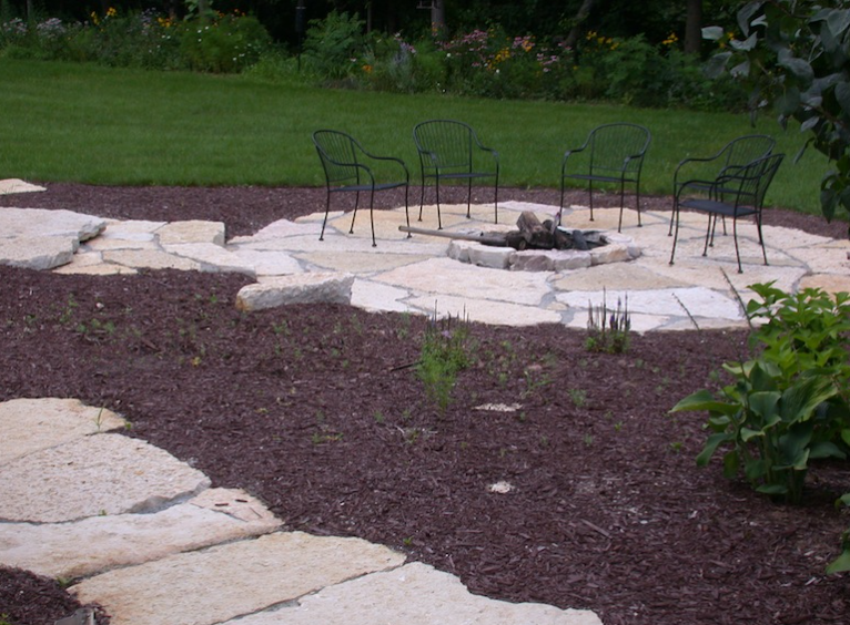 Backyard fire pit surrounded by outdoor chairs on a stone patio with grass and mulch nearby