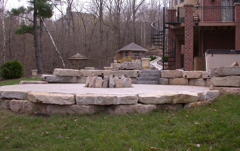 Stone patio with stacked retaining walls and steps in a backyard, bordered by grass and leafless trees