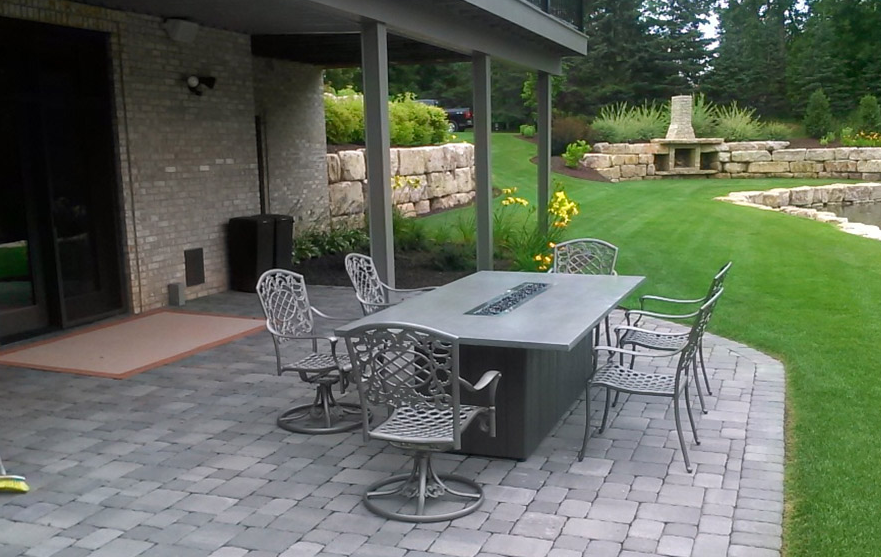 Patio with outdoor dining table and chairs beside a house, overlooking a green lawn and stone fire pit area