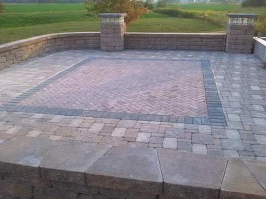 Stone patio with a raised brick-inset center, bordered by low stone walls and pillars overlooking a grassy yard