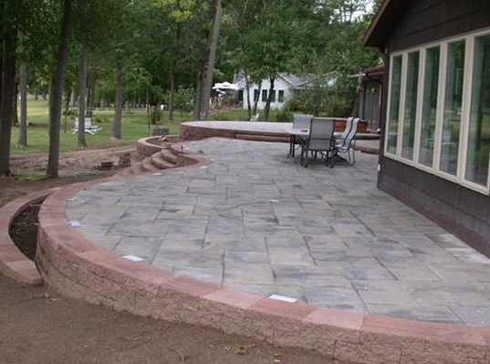 Patio with gray stone pavers, curved brick edging, trees, and outdoor chairs beside a building