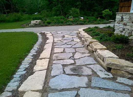 Stone walkway with irregular paving stones bordered by grass and stacked stone edging in a garden.