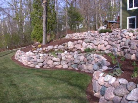 Stone retaining wall terraced into a grassy yard beside trees and a house
