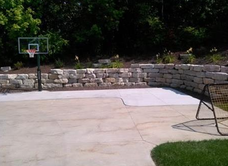 Backyard basketball half-court with hoop, stone retaining wall, and folding chair by a patio