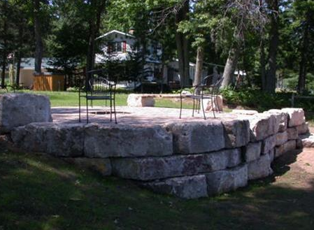 Stone fire pit with black metal grill in a grassy backyard surrounded by trees and houses