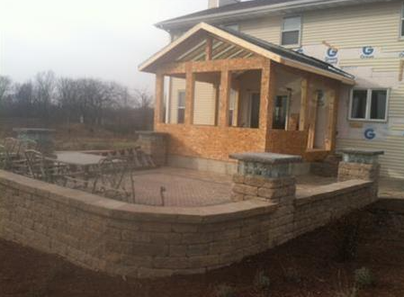 Small brick patio with stone wall and covered porch beside a house in an overcast yard