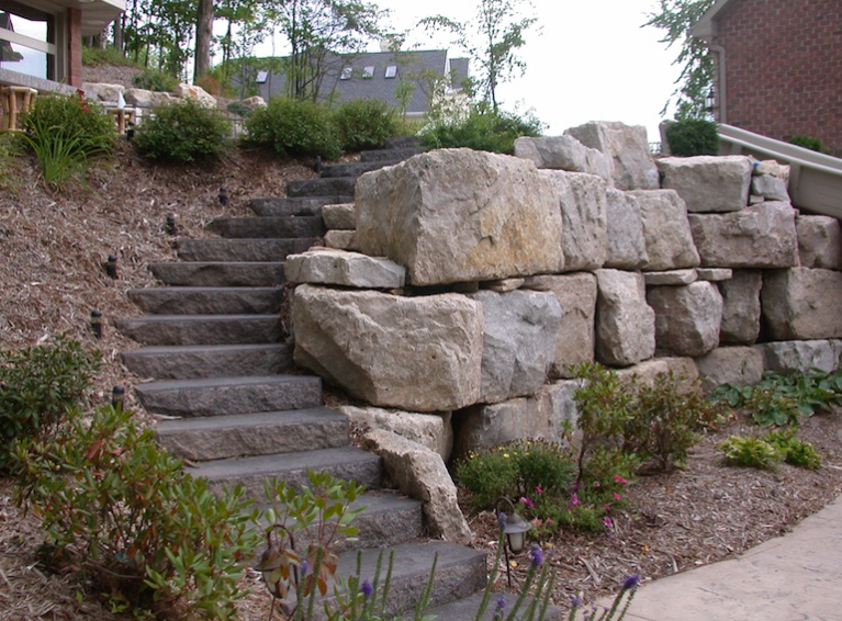 Stone steps beside a large stacked rock retaining wall and landscaped shrubs on a hillside.