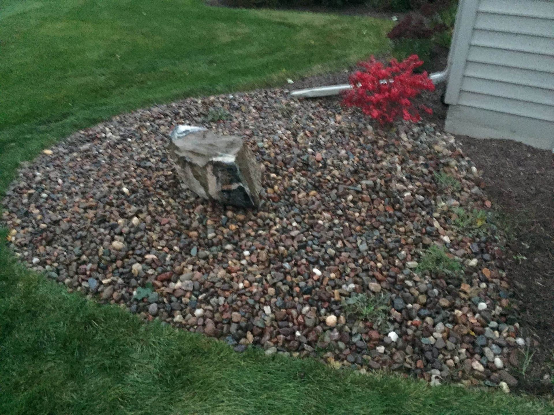 Rock garden with a tree stump centerpiece, small red shrub, and house siding beside a grassy lawn.