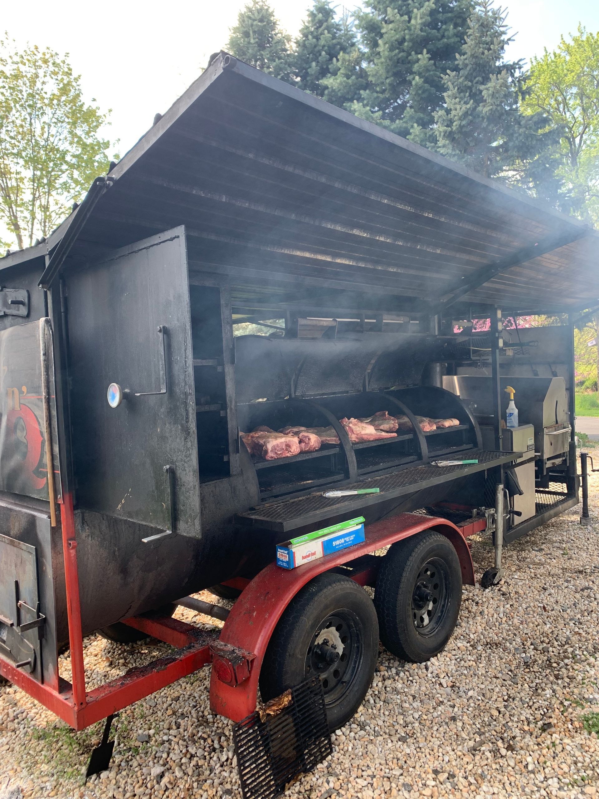 BBQ smoker on a trailer with ribs cooking. Black smoker, red trailer, grey gravel setting.