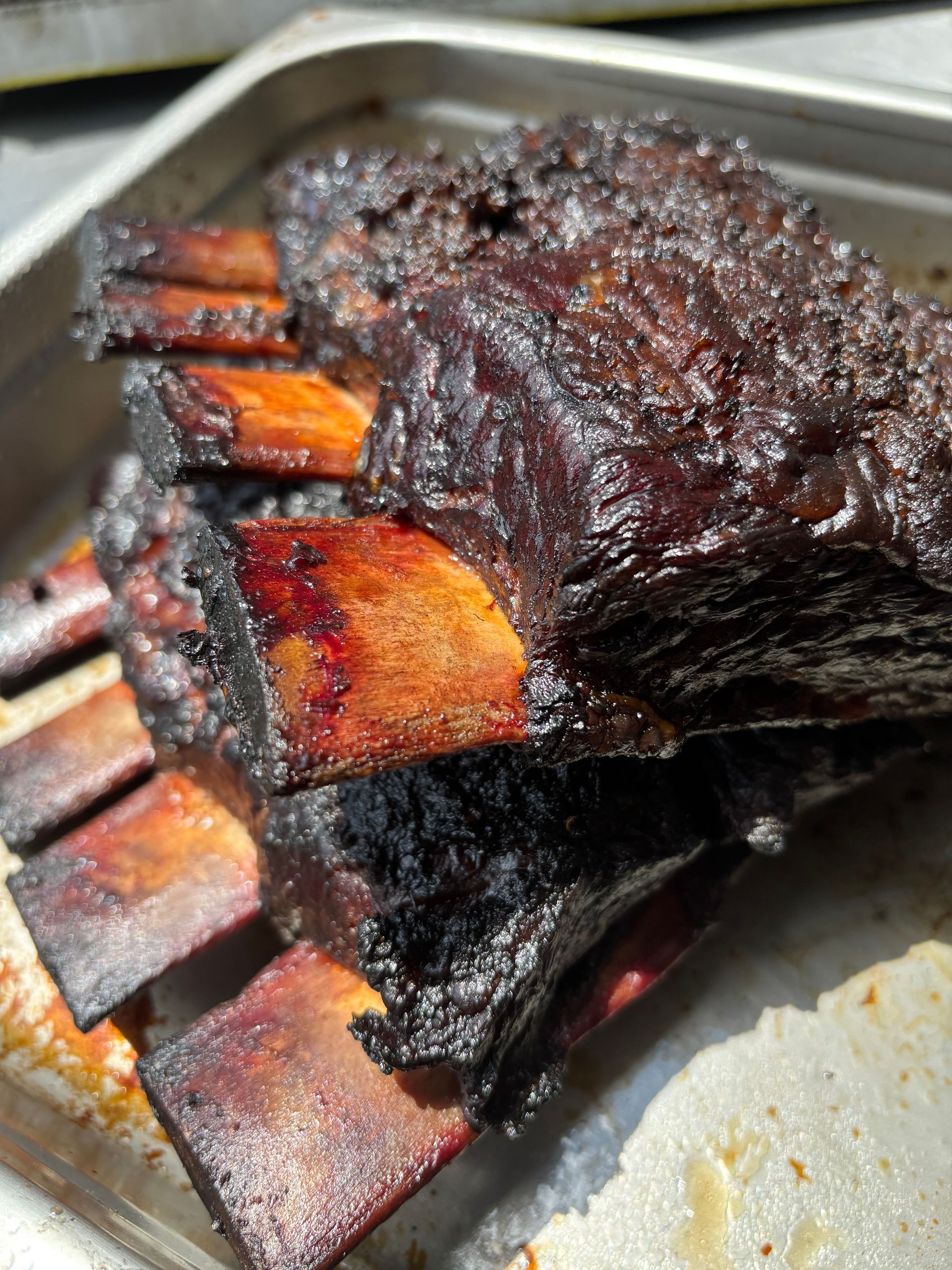 Smoked beef short ribs with charred bark, stacked in a metal tray.
